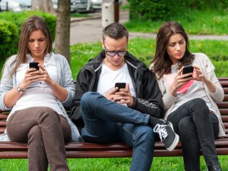 Three young men and women sitting on park bench looking at their phones